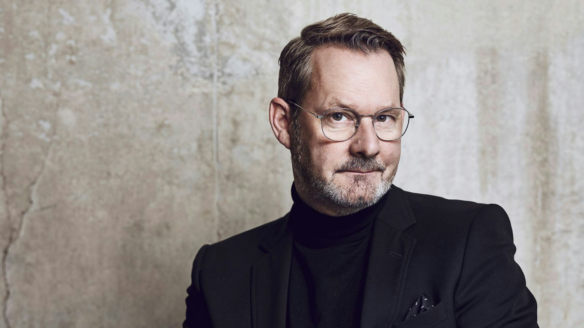 Der Autor Andres Storm sitzt im schwarzen Anzug vor einer Leinwand.