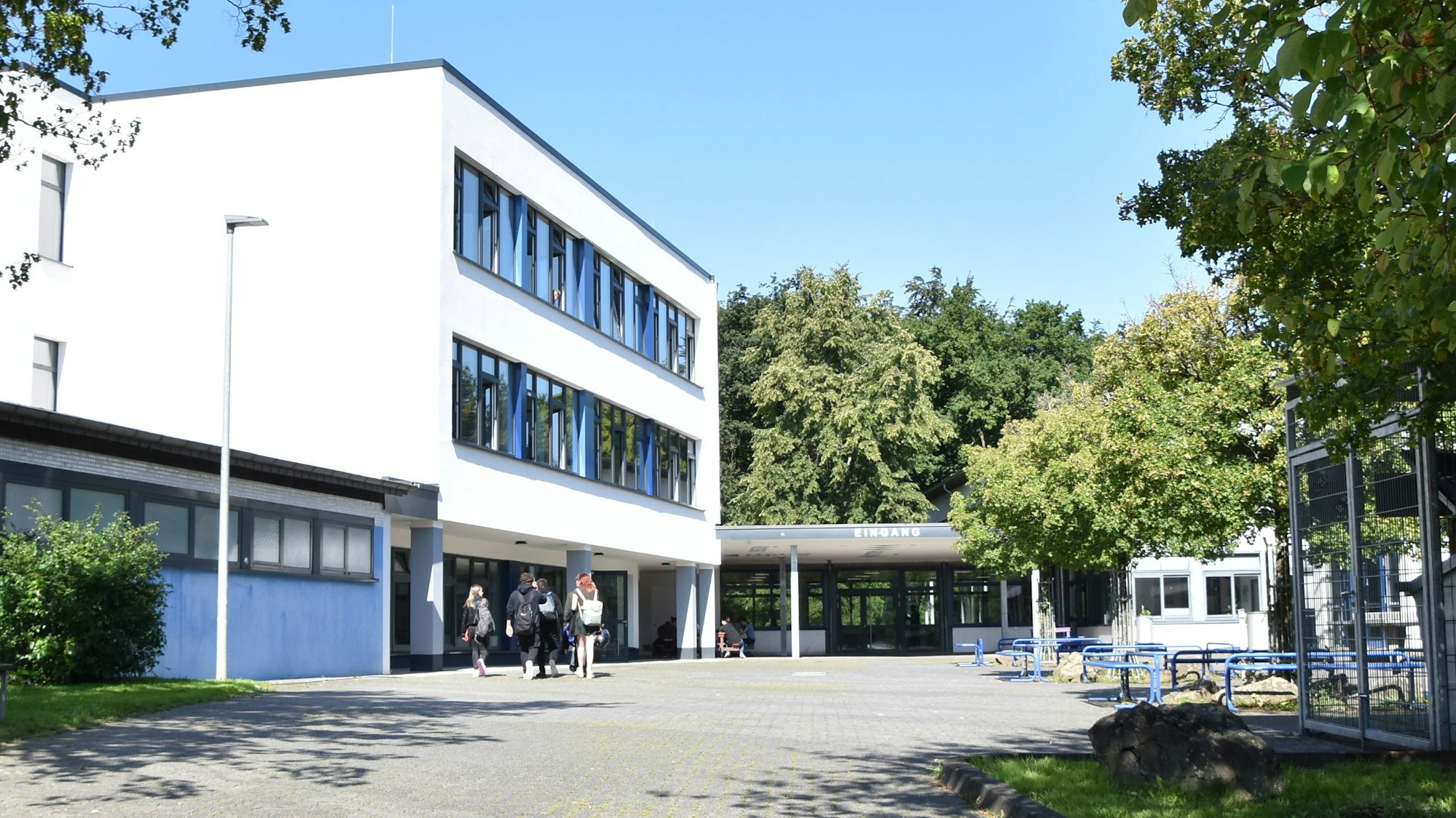 Ein weißes Gebäude, umgeben von Bäumen. Schüler gehen in Richtung des Eingangs.