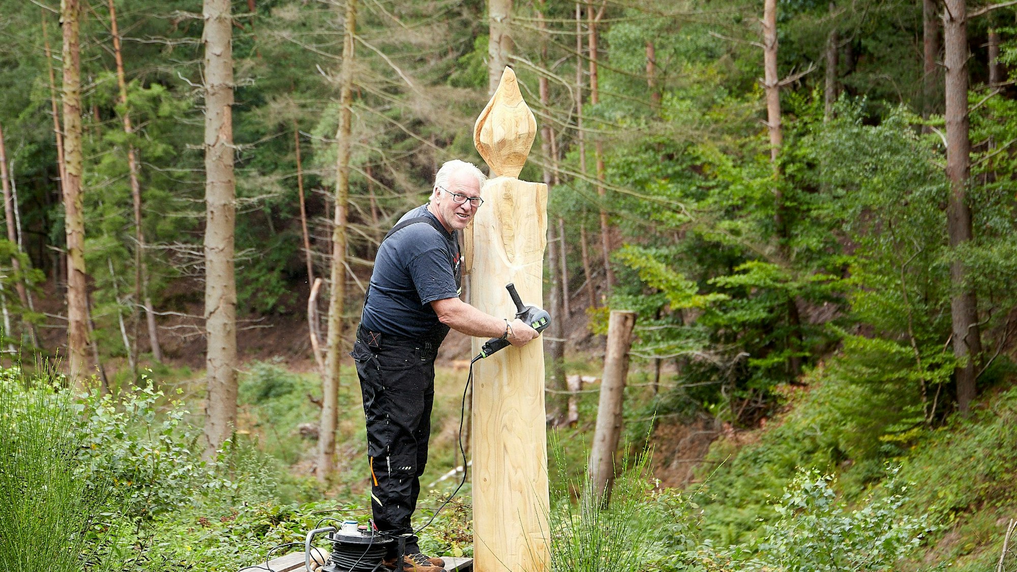 Volker Viktor aus Heimbach steht im Wald neben einer Holzfigur aus einem Baumstamm. Sie hat bereits die Form einer Kerze angenommen.