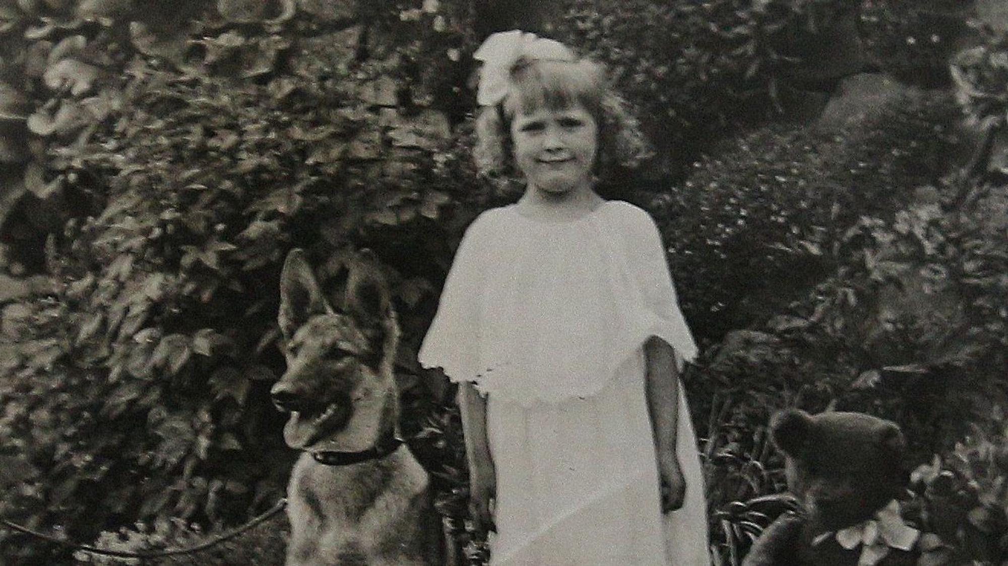Ein Schäferhund und ein Mädchen im weißen Kleid auf einem alten Schwarzweißfoto.