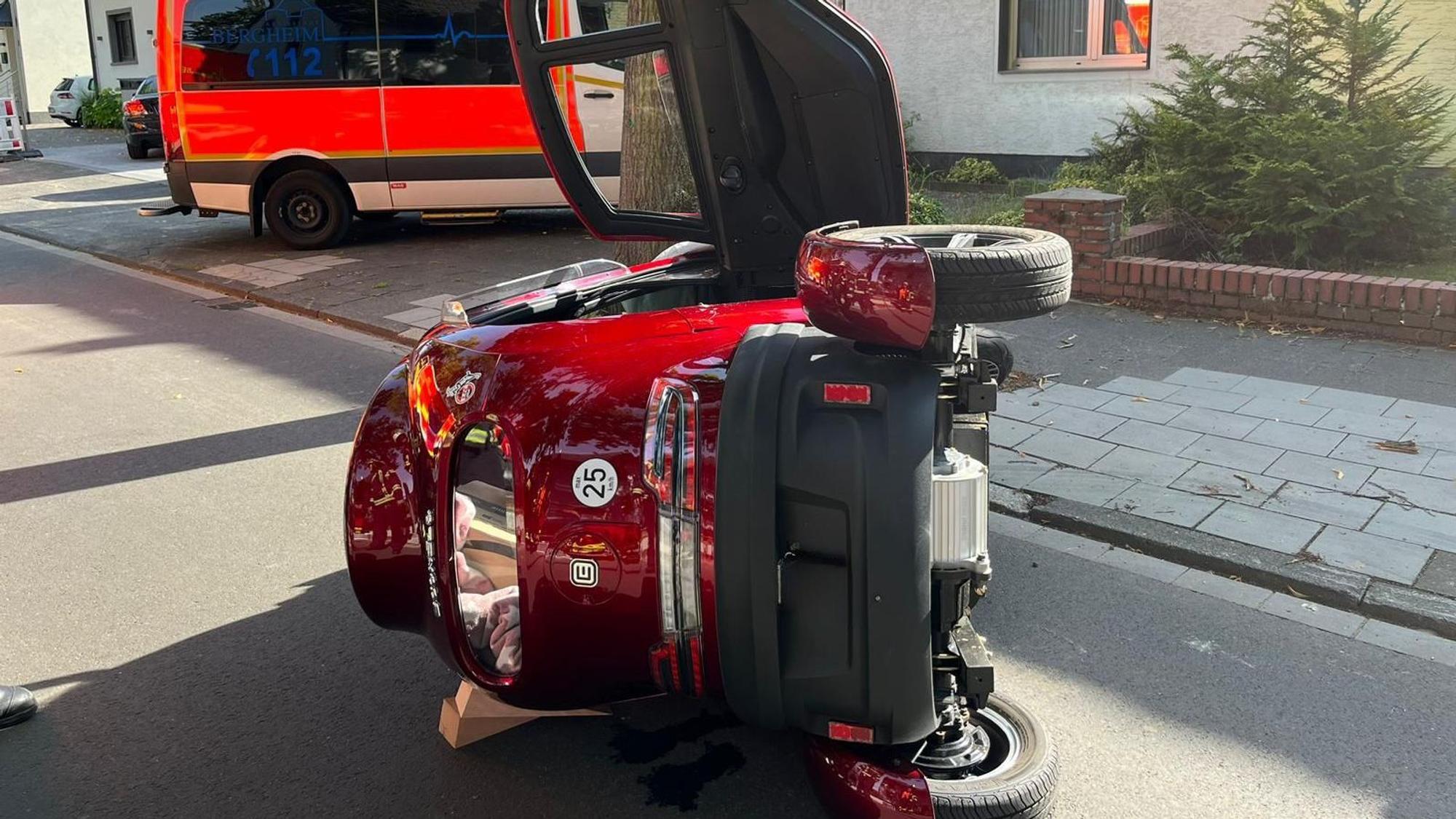 Der weinrote Kabinenroller liegt umgekippt auf der Straße, im Hintergrund steht ein Krankenwagen.