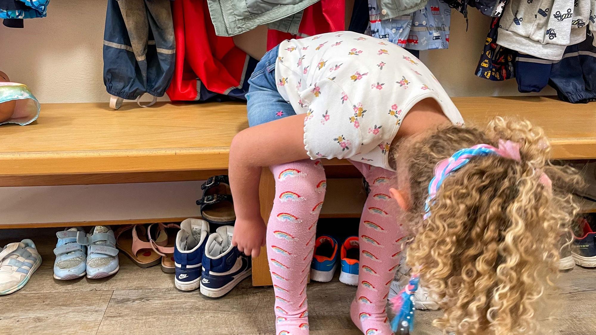 Das Bild zeigt ein Kindergartenkind, das sich auf einer Bank in der Kita die Schuhe anziehen will.