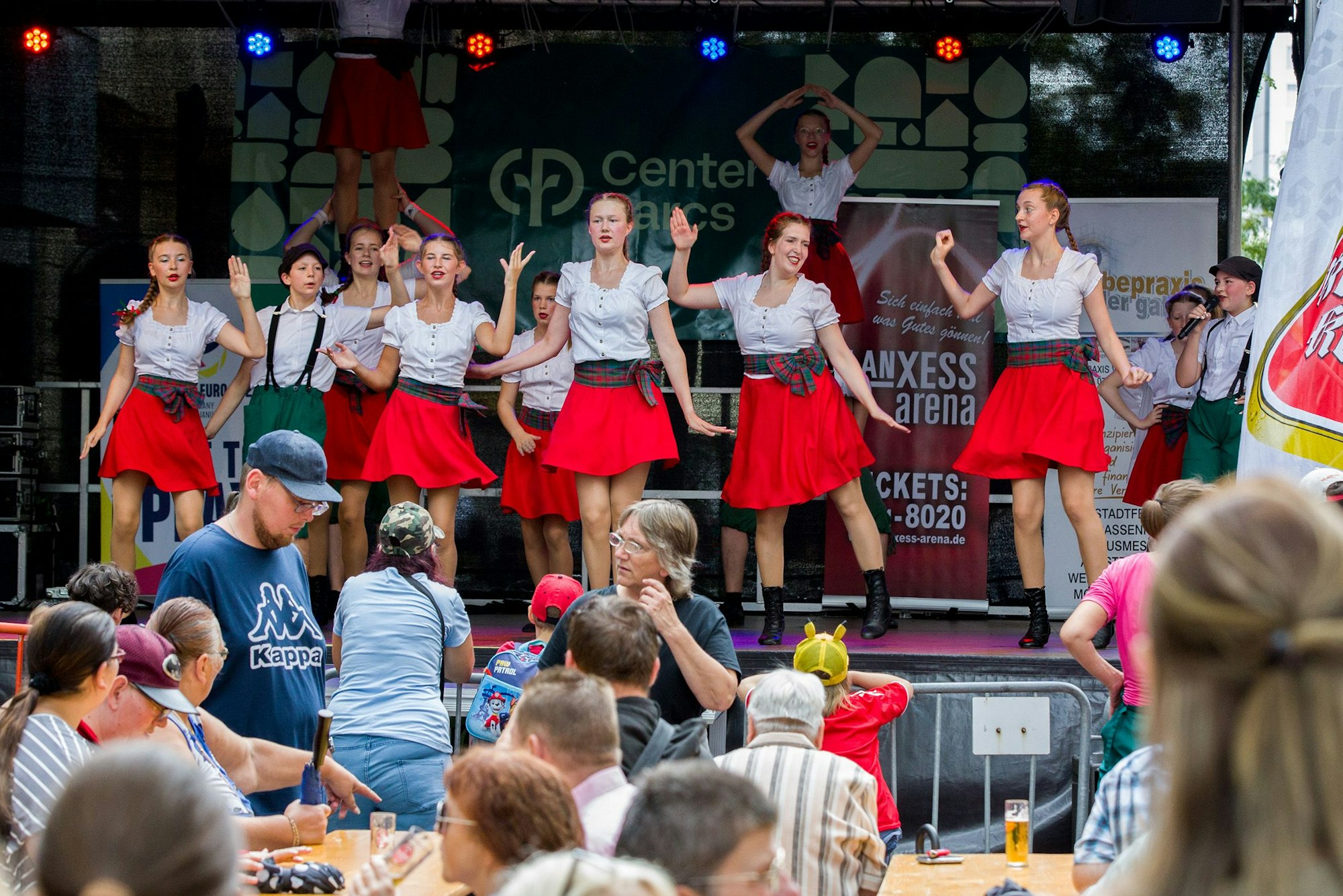 Frauen in weißen Blusen und roten Röcken tanzen auf einer Bühne.