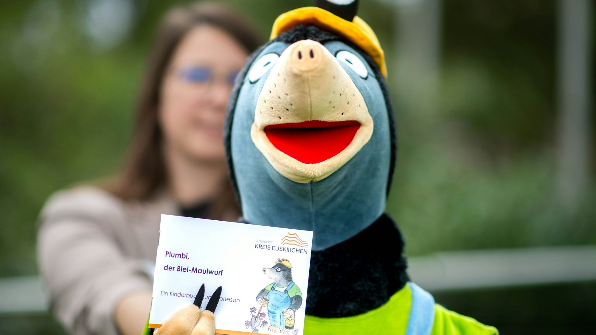 Das Bild zeigt die Handspielfigur Plumbi, die ein kleines Buch in die Kamera hält.