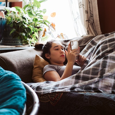 Eine Jugendliche, die unter einer Decke auf der Couch liegt und etwas auf dem Handy sieht.