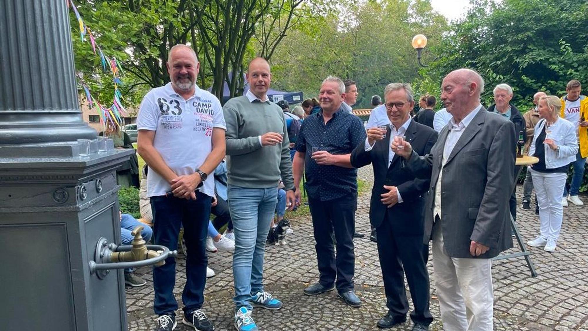 Eine Gruppe von Menschen mit Gläsern in der Hand neben der Pumpe.
