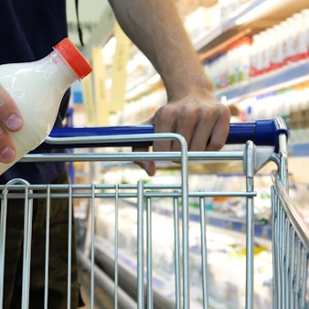 Nahaufnahme einer männlichen Käuferhand, die eine Plastikflasche Milch in einen Einkaufswagen legt.