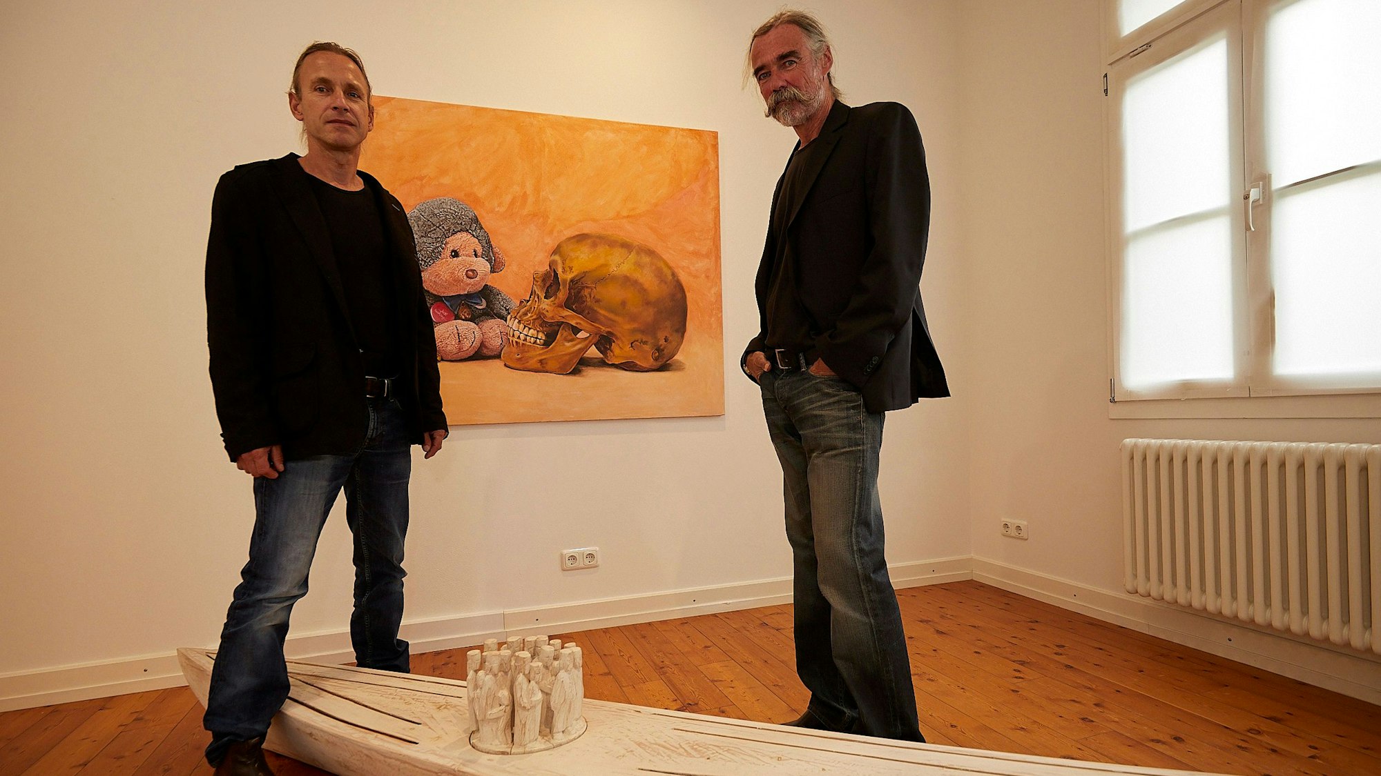Götz Sambale (l.) und Fabian Hochscheid stehen neben ihrer Kunst: einem Gemälde, das ein Kuscheltier und einen menschlichen Schädel zeigt, und einem aus Holz geschnitzten Kajak.