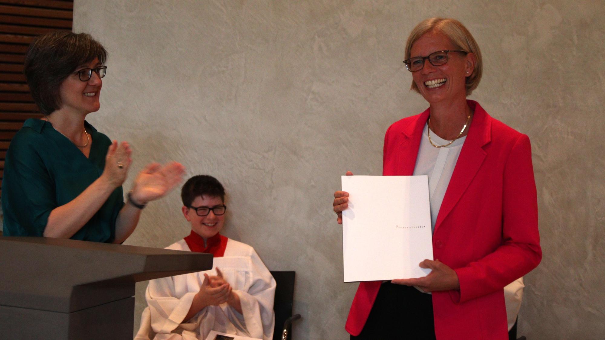 Schulleiterin Carolin Neswadba hält ihre Urkunde, Bernadette Schwarz-Boenneke applaudiert. Ein Messdiener im Hintergrund freut sich.