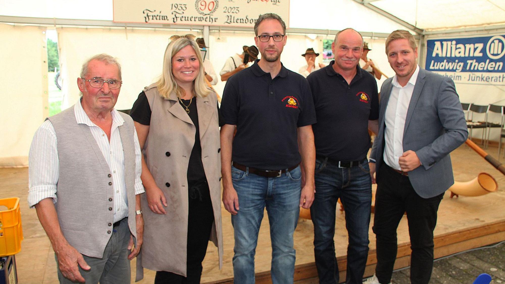 Markus Ramers (r.), Jennifer Meuren und Fritz Reifferscheidt (l.) gratulierten Löschgruppenführer Andreas Palms (M.) und seinem Stellvertreter Martin Bales zum Jubiläum der Löschgruppe. Sie stehen vor einer Bühne in einem Festzelt.