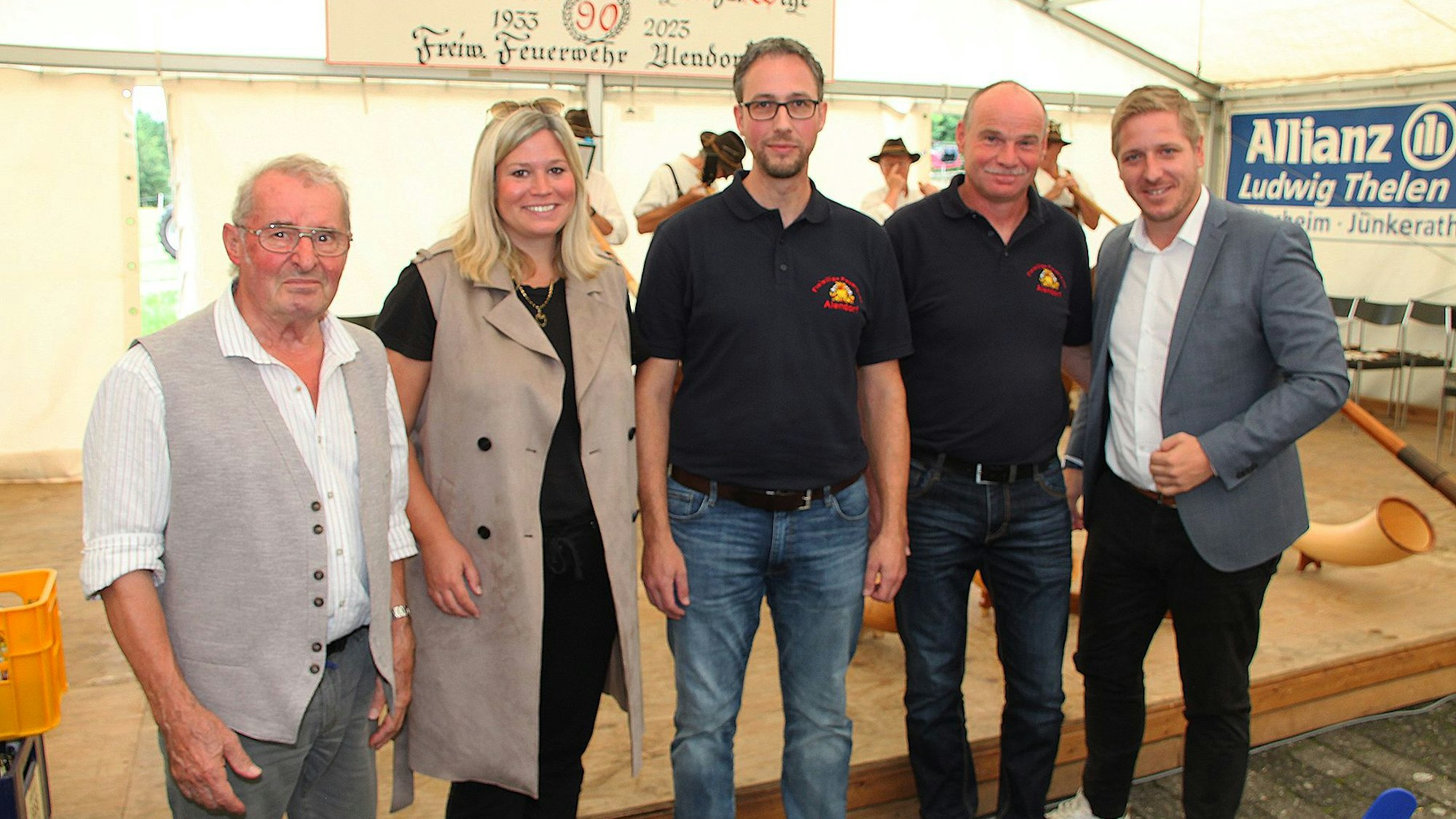 Markus Ramers (r.), Jennifer Meuren und Fritz Reifferscheidt (l.) gratulierten Löschgruppenführer Andreas Palms (M.) und seinem Stellvertreter Martin Bales zum Jubiläum der Löschgruppe. Sie stehen vor einer Bühne in einem Festzelt.