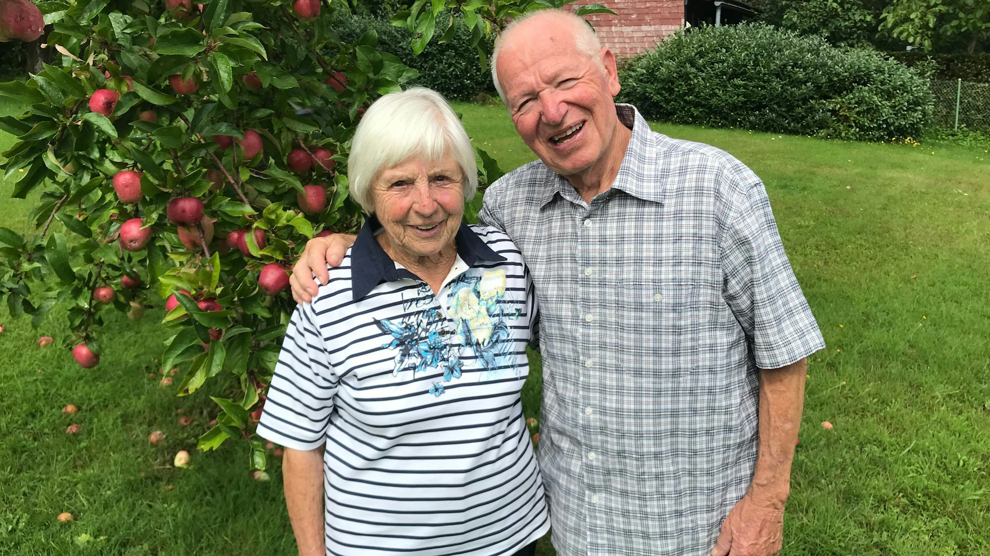 Ein älteres Ehepaar steht Arm in Arm vor einem Apfelbaum.