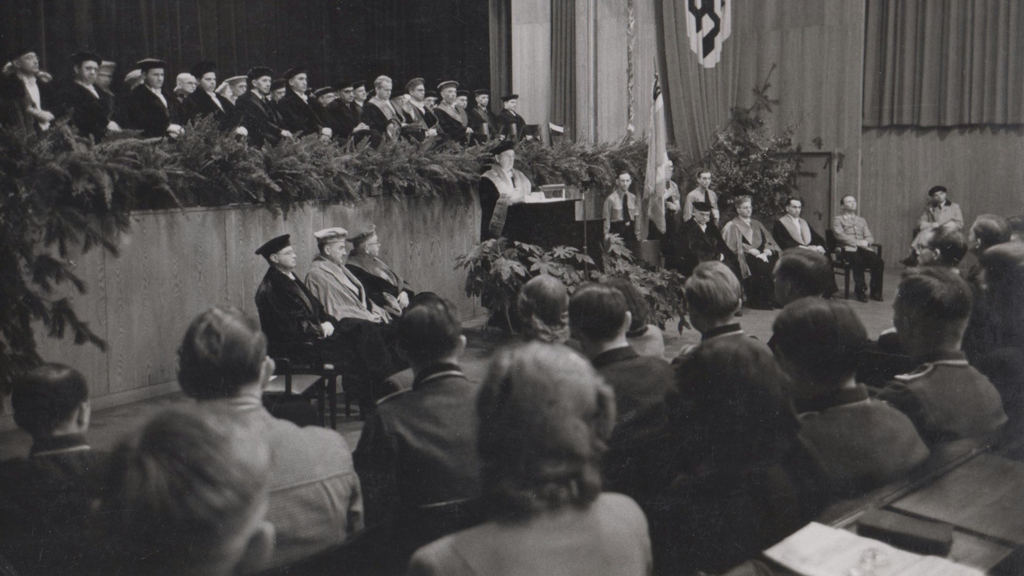 Personen sitzen im Publikum, ein Mann steht an einem Rednerpult und einige Männer sitzen auf einer Bühne und einem Podest.