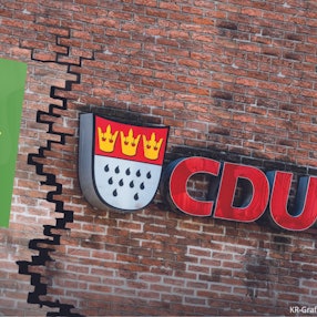 Die Fotomontage zeigt eine Backsteinwand, die einen Riss hat. Auf je einer Seite hängt das Logo von den Grünen und der CDU.