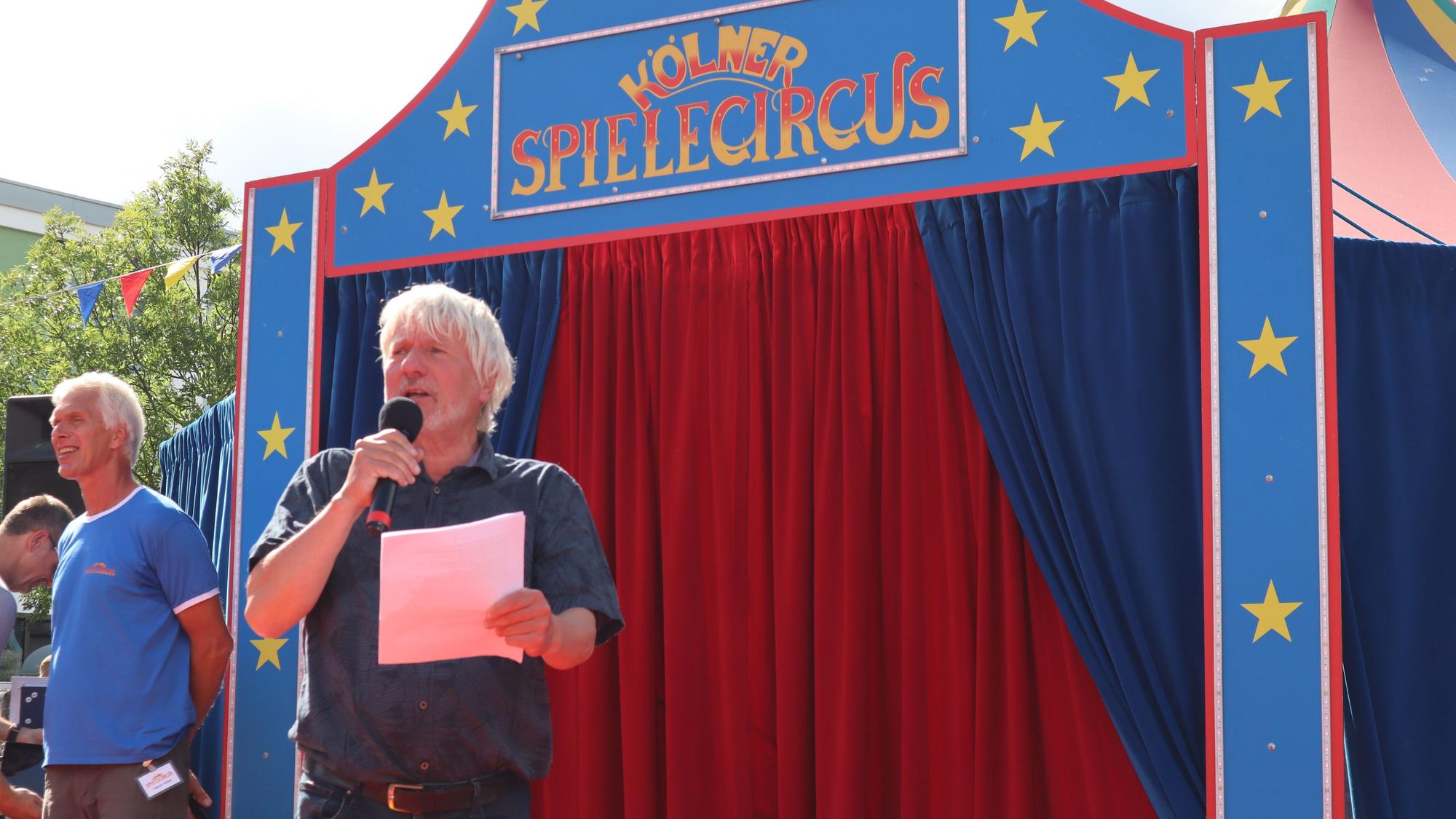 Ein Mann mit grauem Haar und Bart spricht in ein Mikro. Hinter ihm ist der Eingang in die Manege des Kölner Spielecircus.