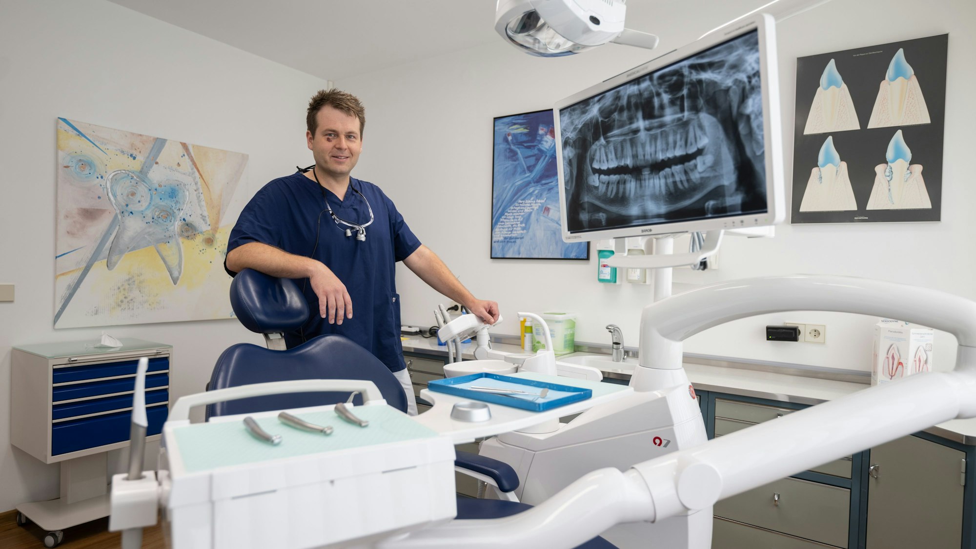 Ein Mann im Ärztekittels lehnt sich an einen Behandlungsstuhl in einem Praxisraum - im Hintergrund hängen Bilder an der Wand.