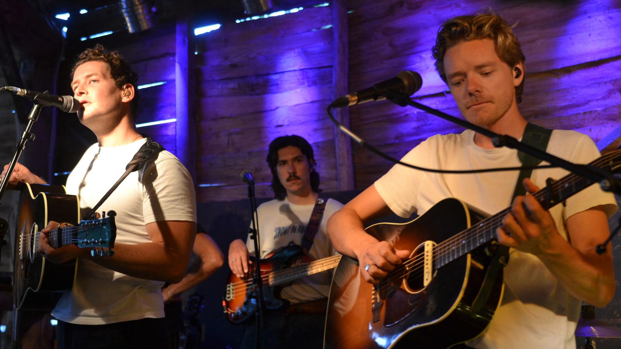 Drei Musiker sitzen auf der Bühne vor jeweils einem Mikrofon. Sie alle spielen Gitarre.