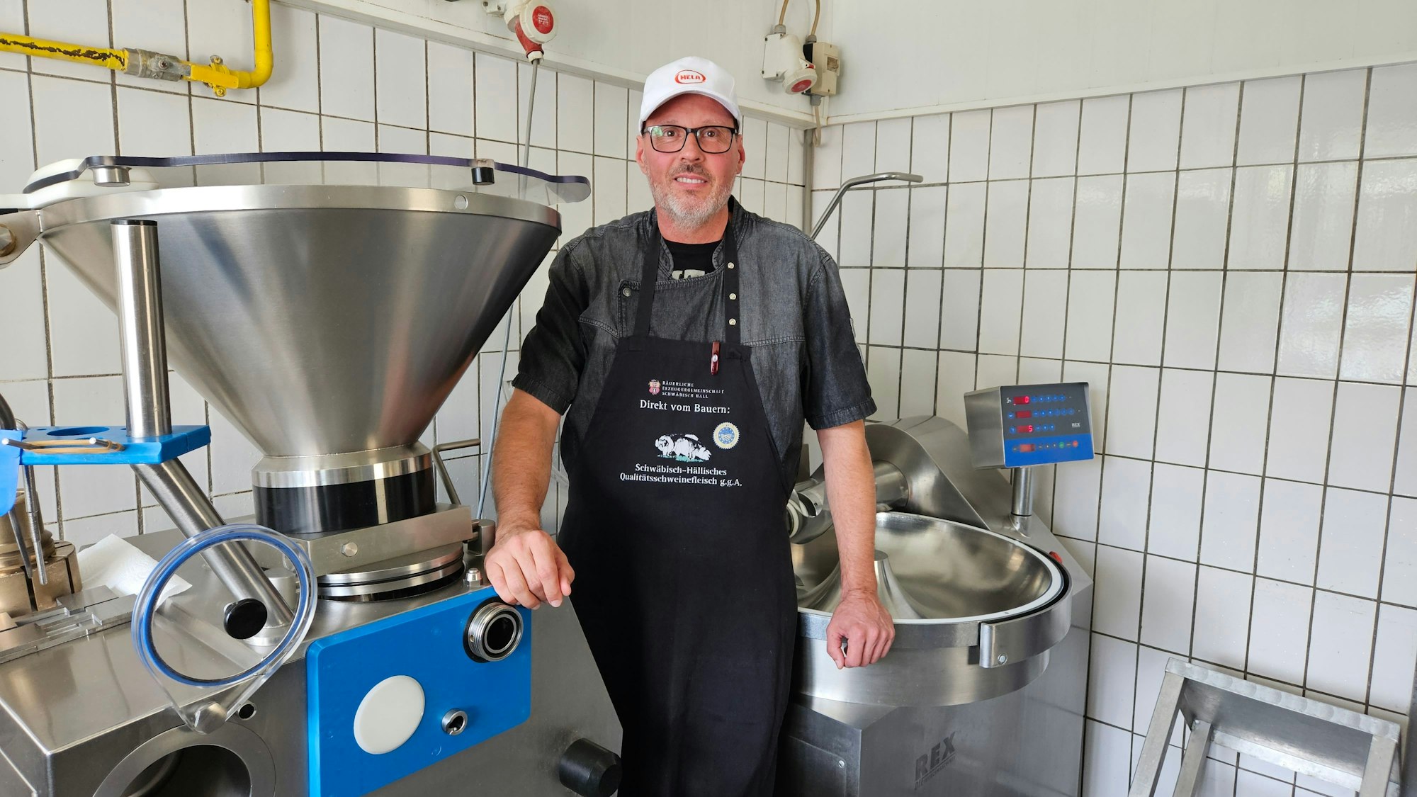 Ein Mann mit Schürze und Kappe steht inmitten von Metzgermaschinen aus silberfarbenem Metall, die Wände des Raums sind gefliest.