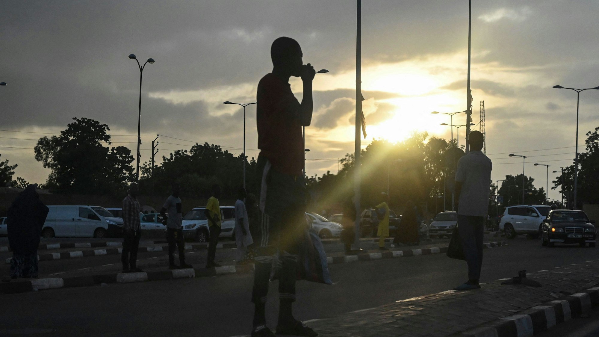 Die Umrisse von Menschen auf einer Straße in Niamey in Niger ist zu sehen - im Hintergrund bricht die Sonne durch eine dichte Wolkendecke.