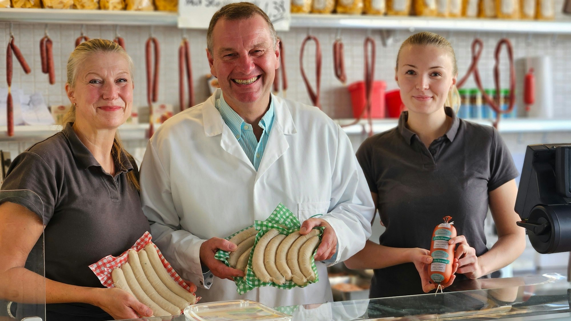 Zwei Frauen und ein Mann stehen hinter der gläsernen Theke einer Metzgerei und zeigen Wurstwaren.