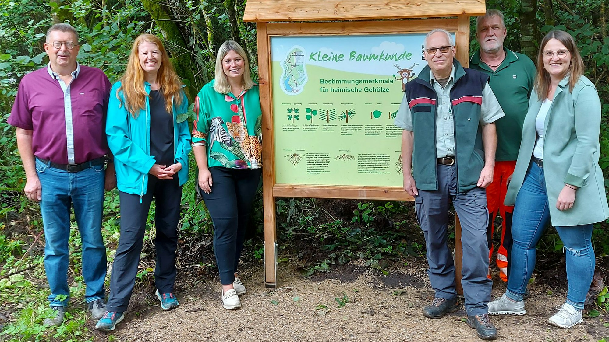 Drei Männer und drei Frauen stehen in Freizeitkleidung im Wald an einem Informationsschild, auf dem „Kleine Baumkunde“ steht. Im Hintergrund sind Bäume und Blätter zu erkennen.