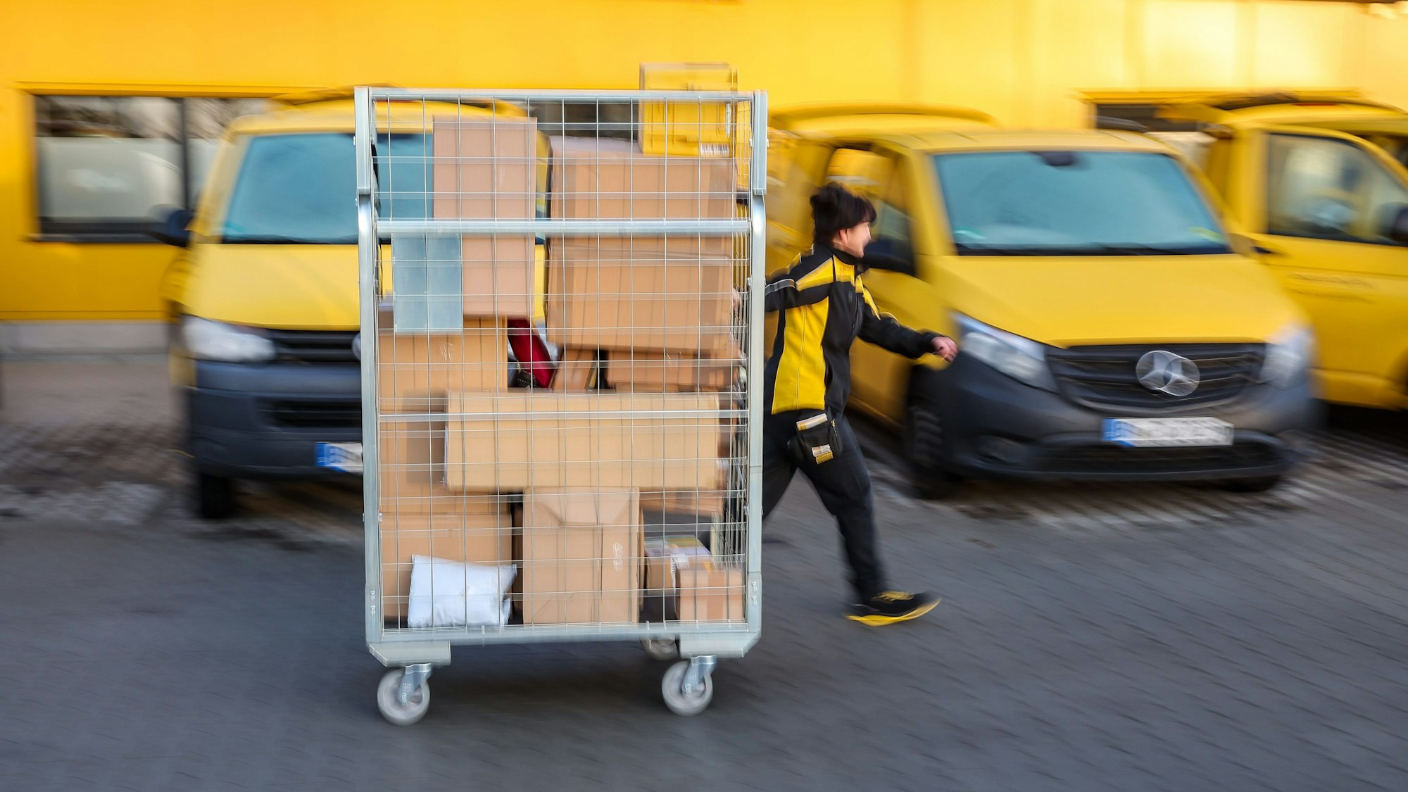 Eine Mitarbeiterin der Deutsche Post DHL geht mit einem vollen Paketwagen zu ihrem Fahrzeug auf dem Hof eines Zustellpunktes.