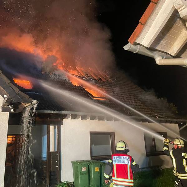 Feuerwehrleute löschen mit Wasser die Flammen, die aus dem Dach eines Hauses schlagen.