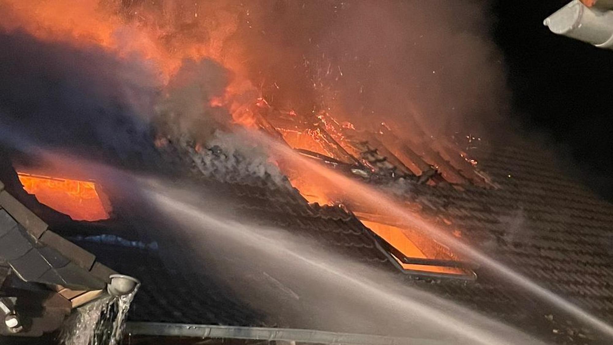 Flammen schlagen aus einem Dach, Wasserstrahle sind auf die Flammen gerichtet.