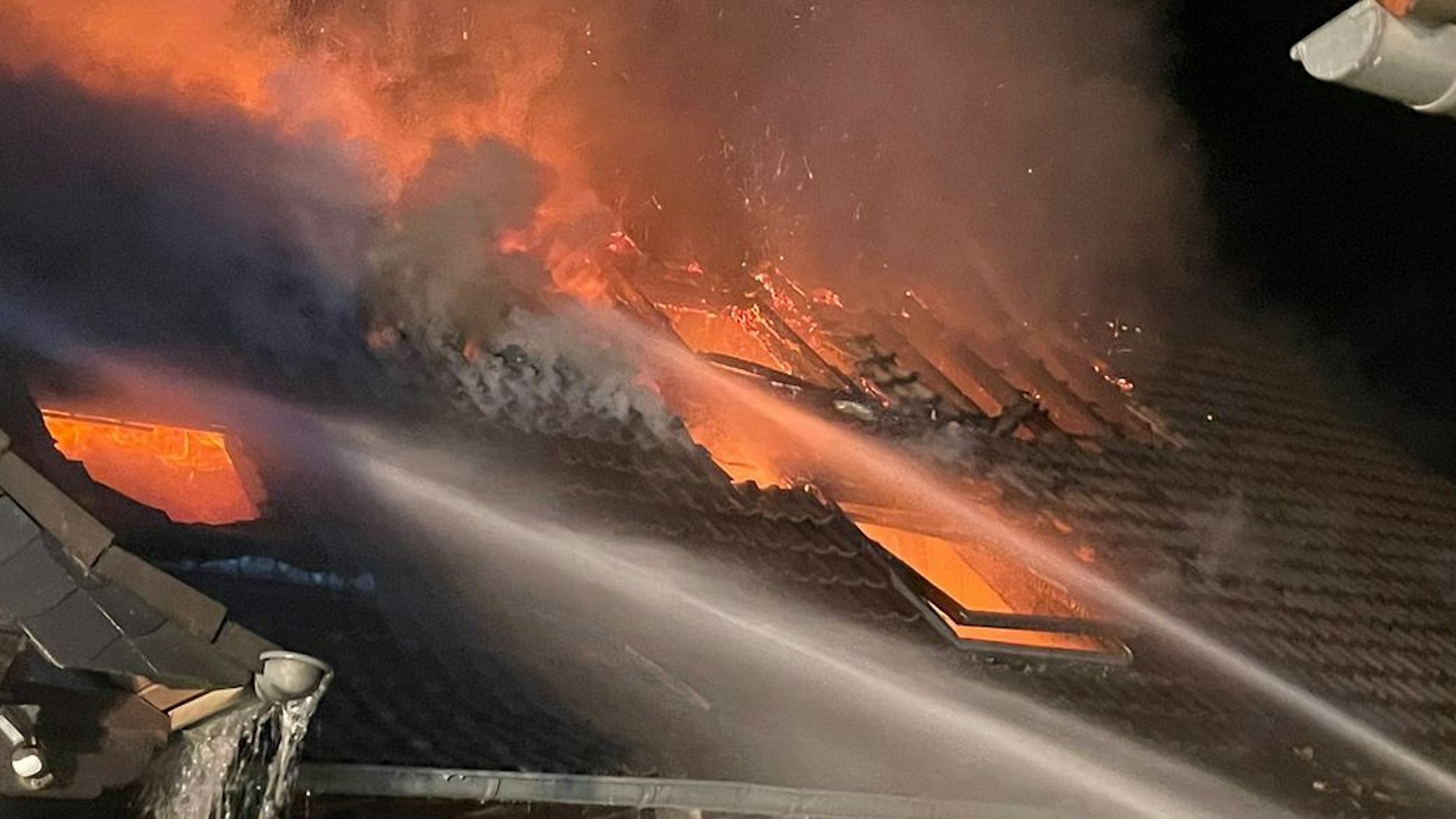 Flammen schlagen aus einem Dach, Wasserstrahle sind auf die Flammen gerichtet.