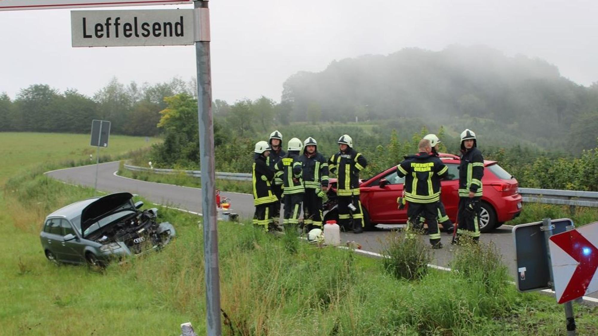 Ein Unfallauto steht auf der Straße, umringt von Feuerwehrleuten. ein weiteres Auto steht mit geöffneter Motorhaube in der Wiese neben der Straße.