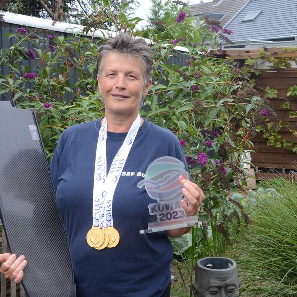 Eine Frau mit kurzen grauen Haaren steht in ihrem Garten. Sie hat drei Goldmedaillen um den Hals hängen; im rechten Arm trägt sie Schwimmflossen. In der linken Hand hält sie eine bunte Trophäe von der Weltmeisterschaft