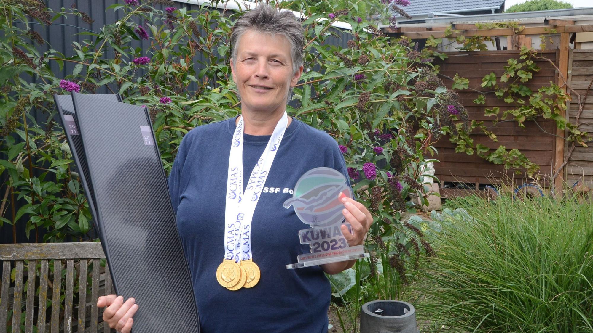 Eine Frau mit kurzen grauen Haaren steht in ihrem Garten. Sie hat drei Goldmedaillen um den Hals hängen; im rechten Arm trägt sie Schwimmflossen. In der linken Hand hält sie eine bunte Trophäe von der Weltmeisterschaft