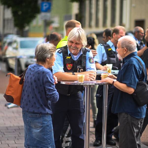 Der Bezirksdienstbeamte Roger Kath steht mit zwei Euskirchener Bürgern an einem Stehtisch. Sie haben jeweils ein Glas mit Kaffee vor sich stehen.