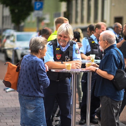 Der Bezirksdienstbeamte Roger Kath steht mit zwei Euskirchener Bürgern an einem Stehtisch. Sie haben jeweils ein Glas mit Kaffee vor sich stehen.