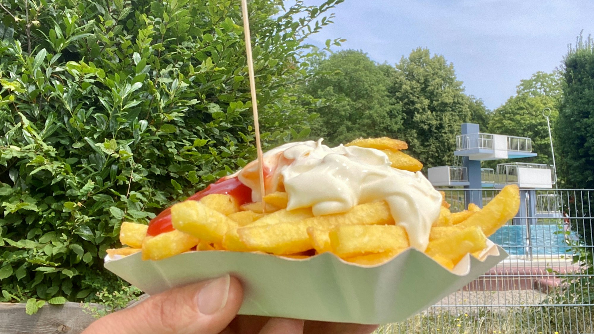 In einer Pappschale liegen Pommes frites mit Ketchup und Mayo sowie einer hölzernen Gabel, im Hintergrund ist ein Sprungturm eines Freibads zu sehen.