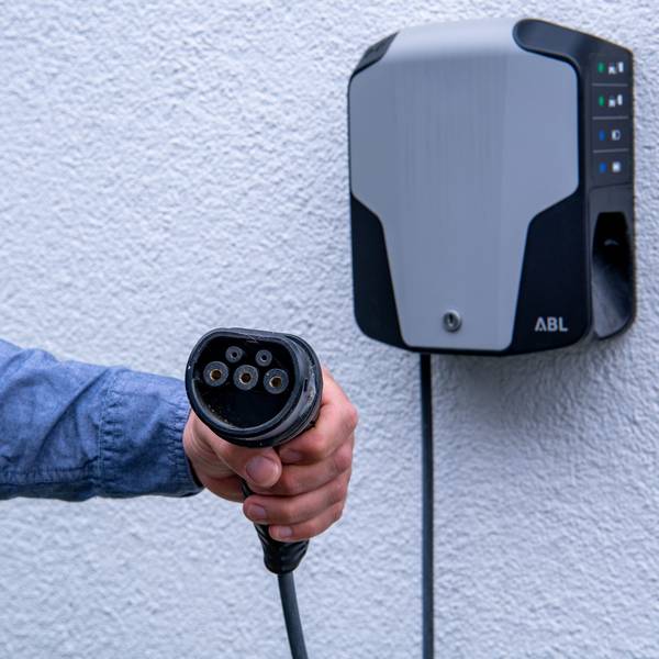 Eine Hand hät enen Anschluss zum Aufladen von E-Autos, im Hintergrund ist eine Wallbox an einer Wand zu sehen.