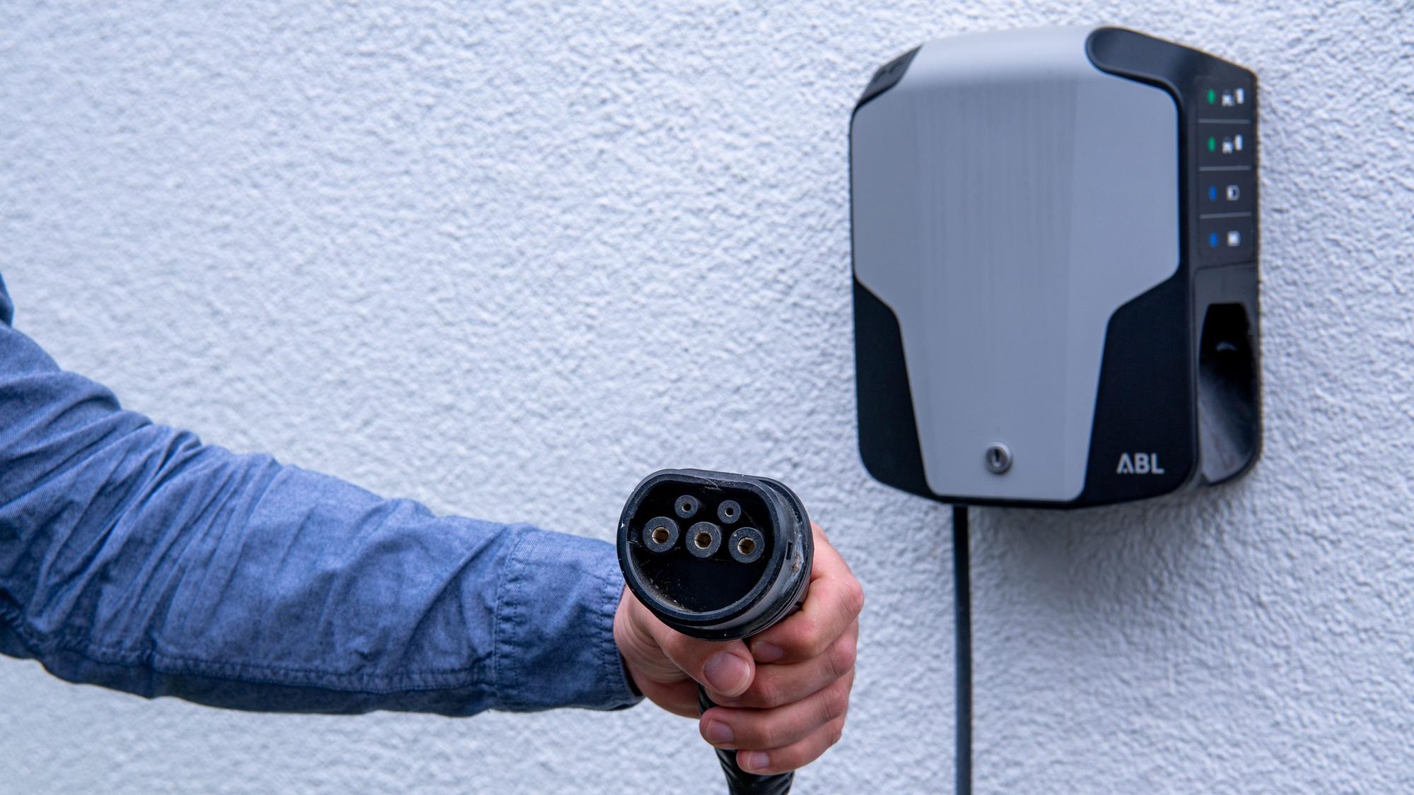 Eine Hand hät enen Anschluss zum Aufladen von E-Autos, im Hintergrund ist eine Wallbox an einer Wand zu sehen.