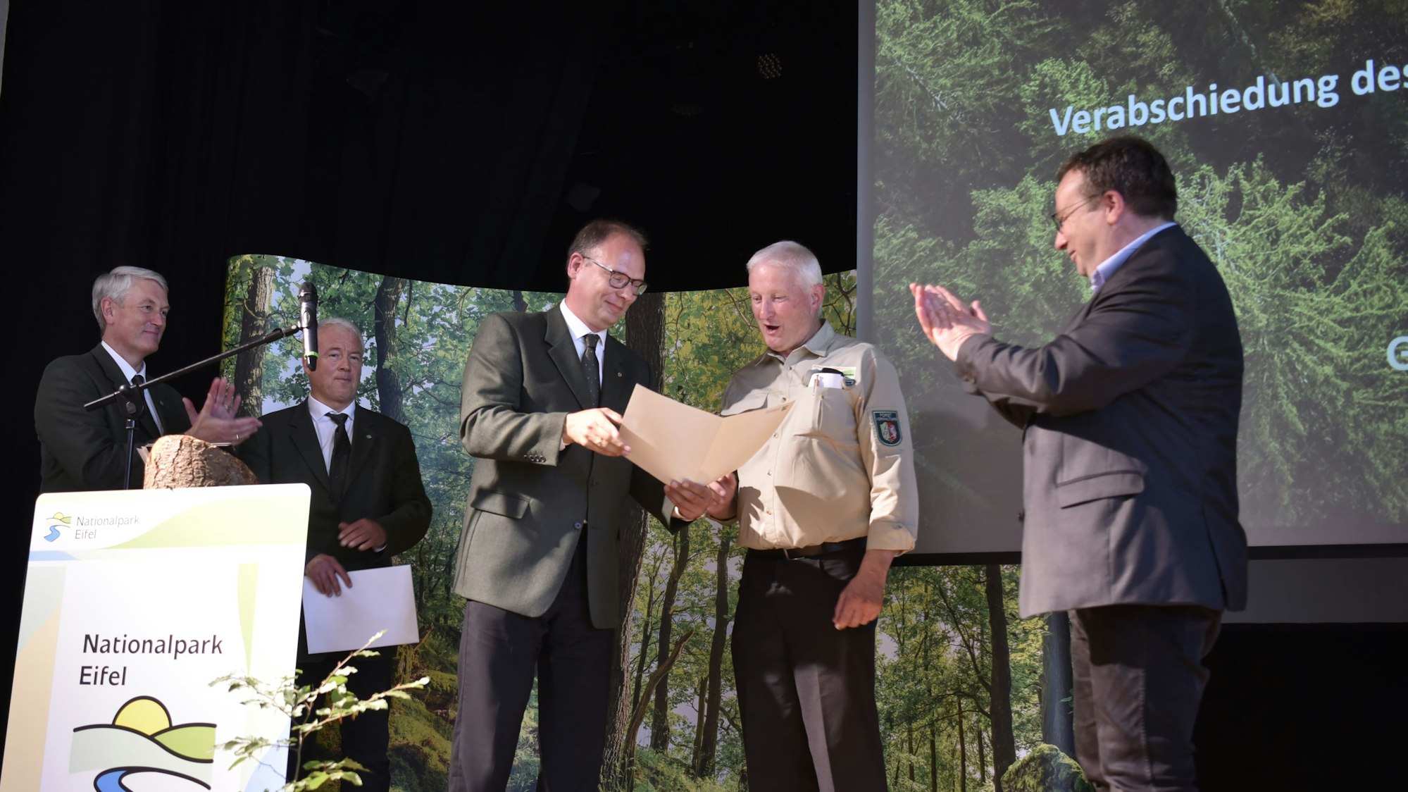 Einige Männer stehen auf einer Bühne. Einer überreicht einem anderen eine Urkunde. Auf einem Schild vorne an der Bühne steht: „Nationalpark Eifel“.