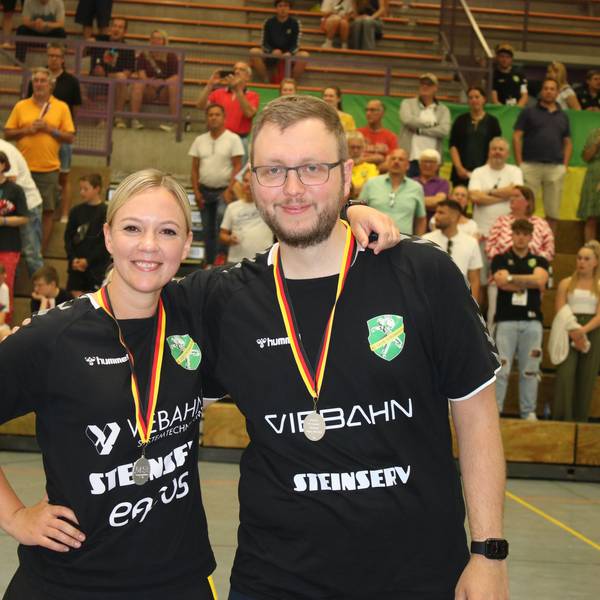 Zwei Trainer, ein Mann und eine Frau, stehen mit Medaillen in einer Handballhalle um den Hals nebeneinander.