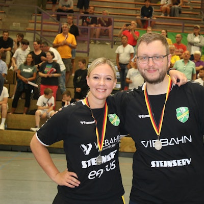 Zwei Trainer, ein Mann und eine Frau, stehen mit Medaillen in einer Handballhalle um den Hals nebeneinander.