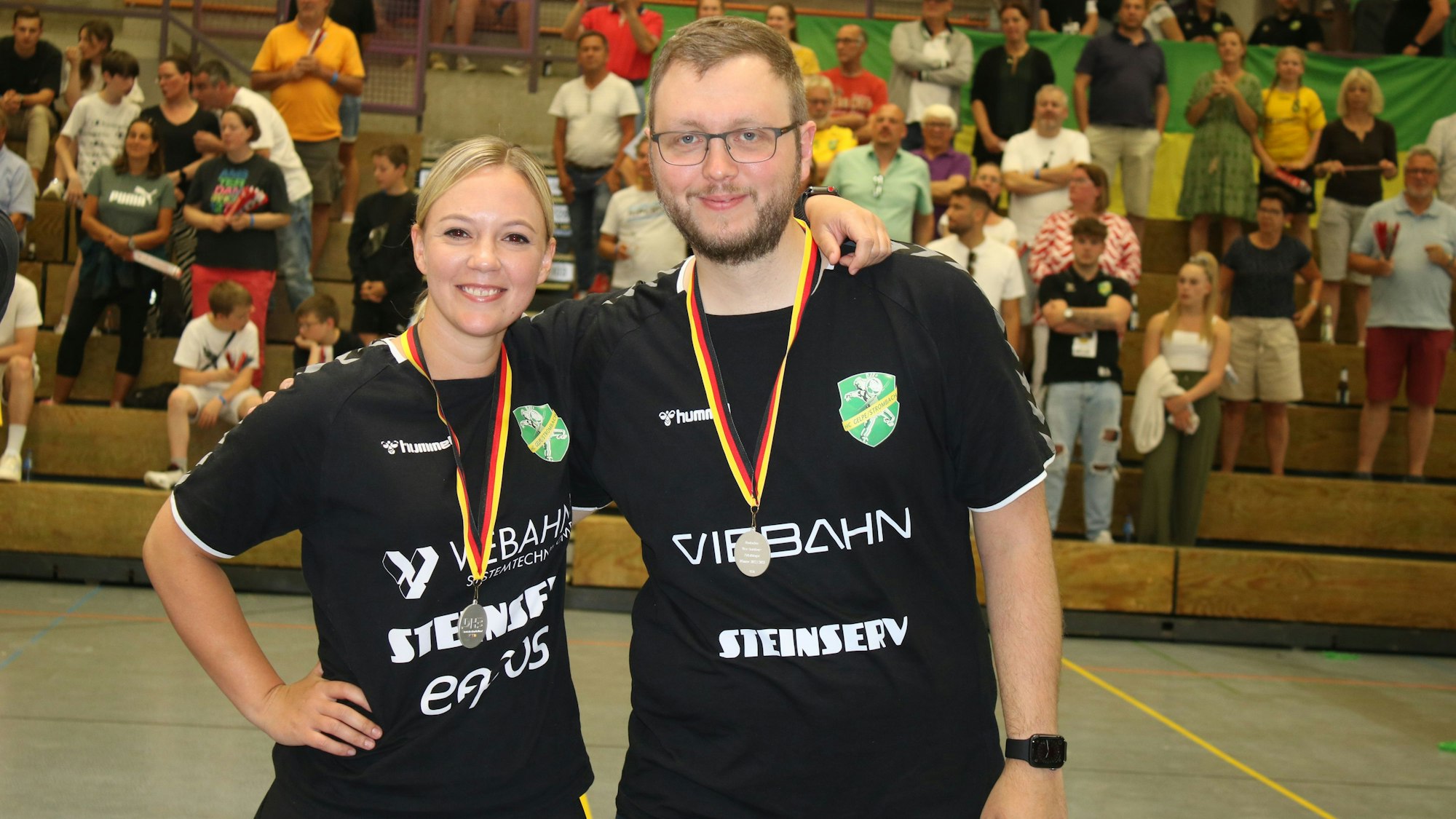 Zwei Trainer, ein Mann und eine Frau, stehen mit Medaillen in einer Handballhalle um den Hals nebeneinander.