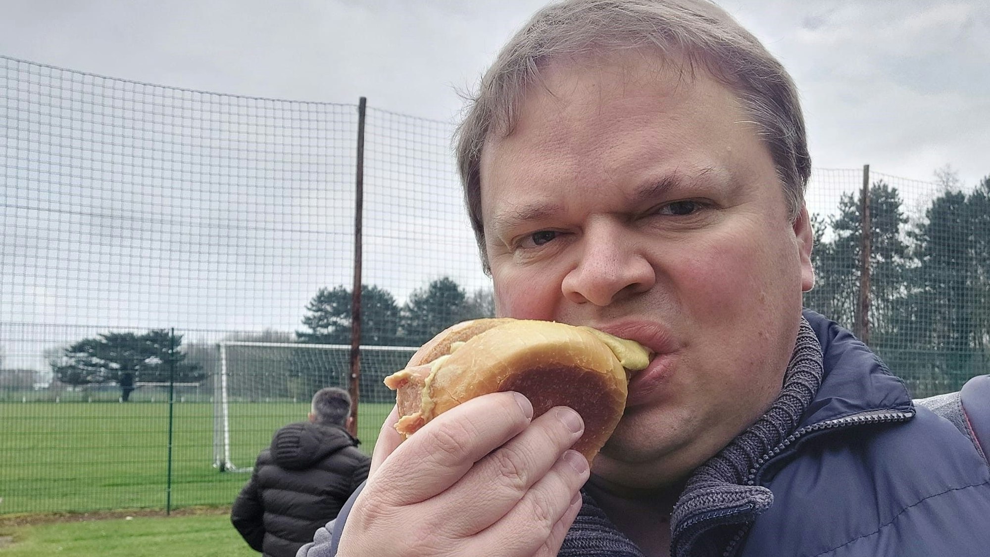 Sportreporter Rocco Bartsch beißt am Rande eines Fußballspiels in ein Bratwurstbrötchen.