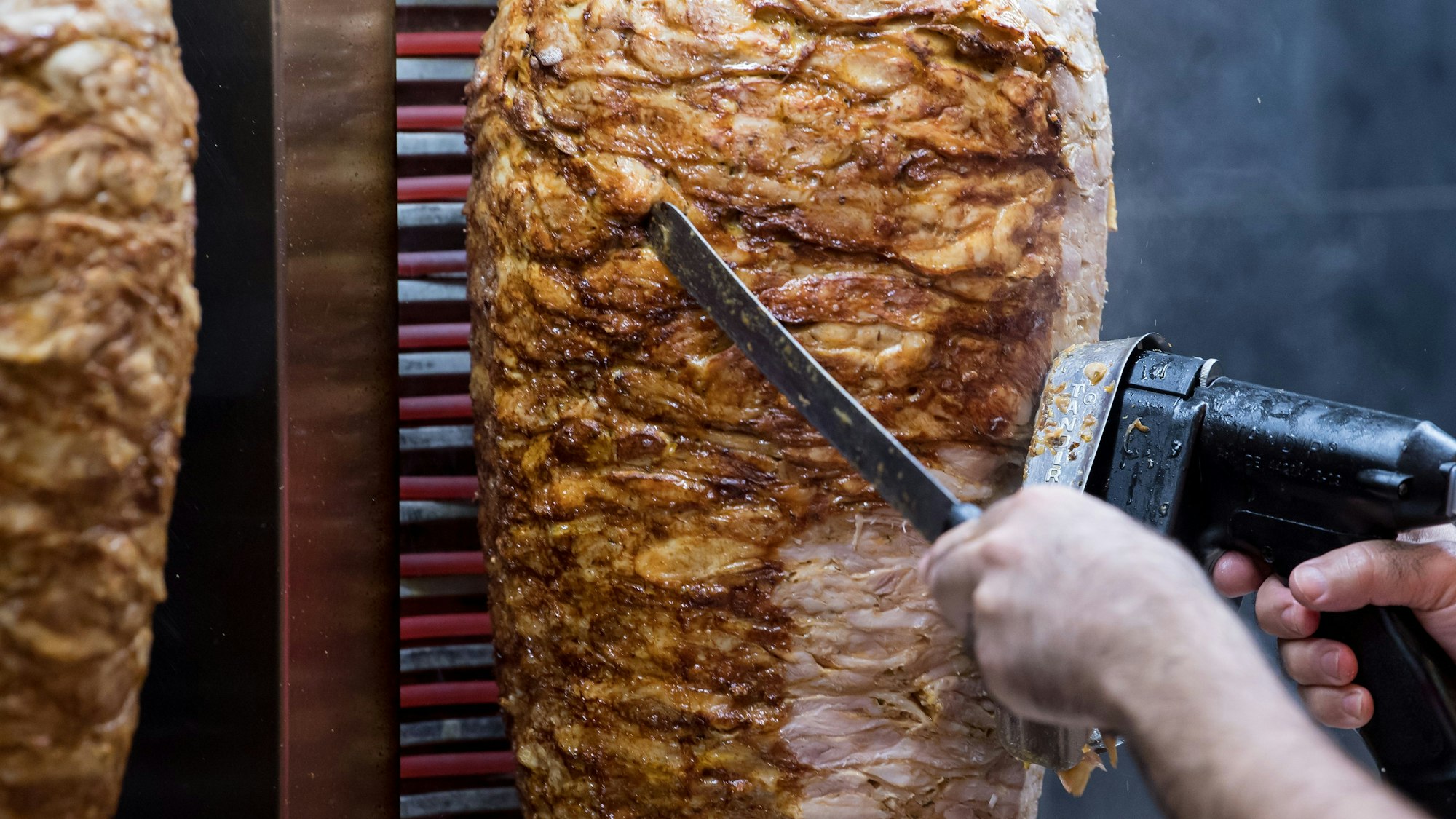 Dönerspieße drehen sich in einem Imbiss. Ein Mitarbeiter schneidet mit einem Werkzeug Fleisch ab. (Symbolbild)