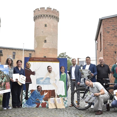 Eine Gruppe von Menschen steht auf einem Platz. Im Hintergrund ist ein mittelalterlicher Turm zu erkennen.