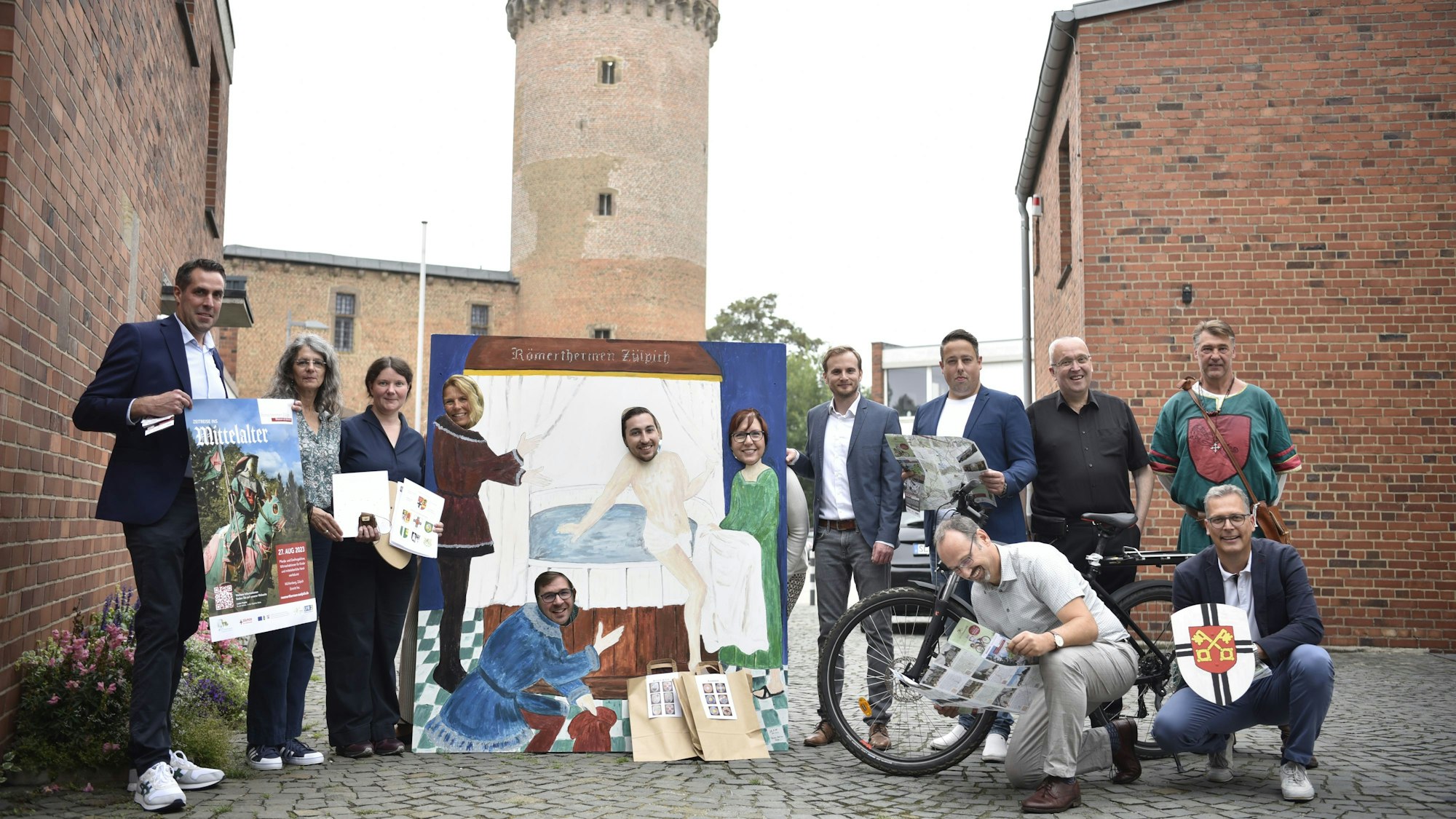 Eine Gruppe von Menschen steht auf einem Platz. Im Hintergrund ist ein mittelalterlicher Turm zu erkennen.
