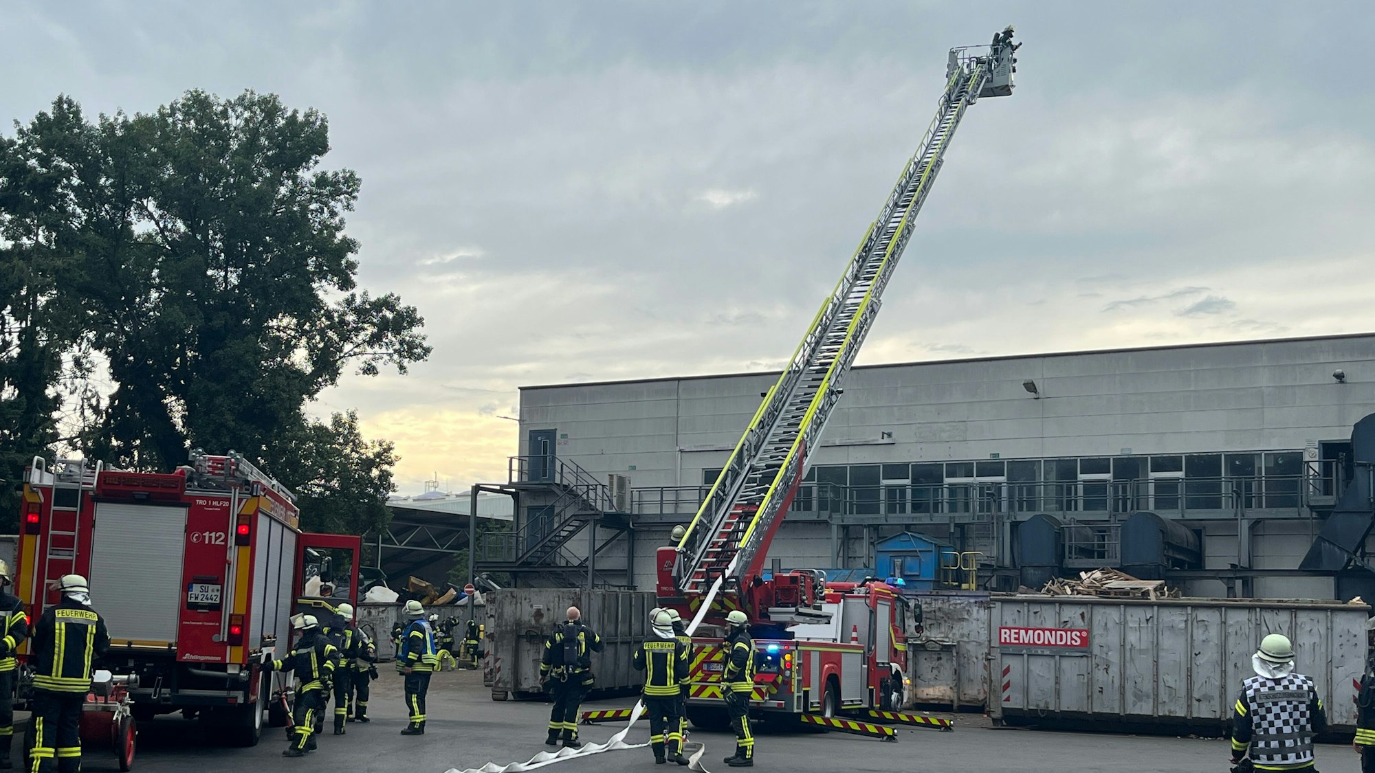 Feuerwehrleute stehen mit mehreren Löschfahrzeugen und einer Drehleiter an einer Industriehalle, Container mit dem Schriftzug Remondis stehen davor.