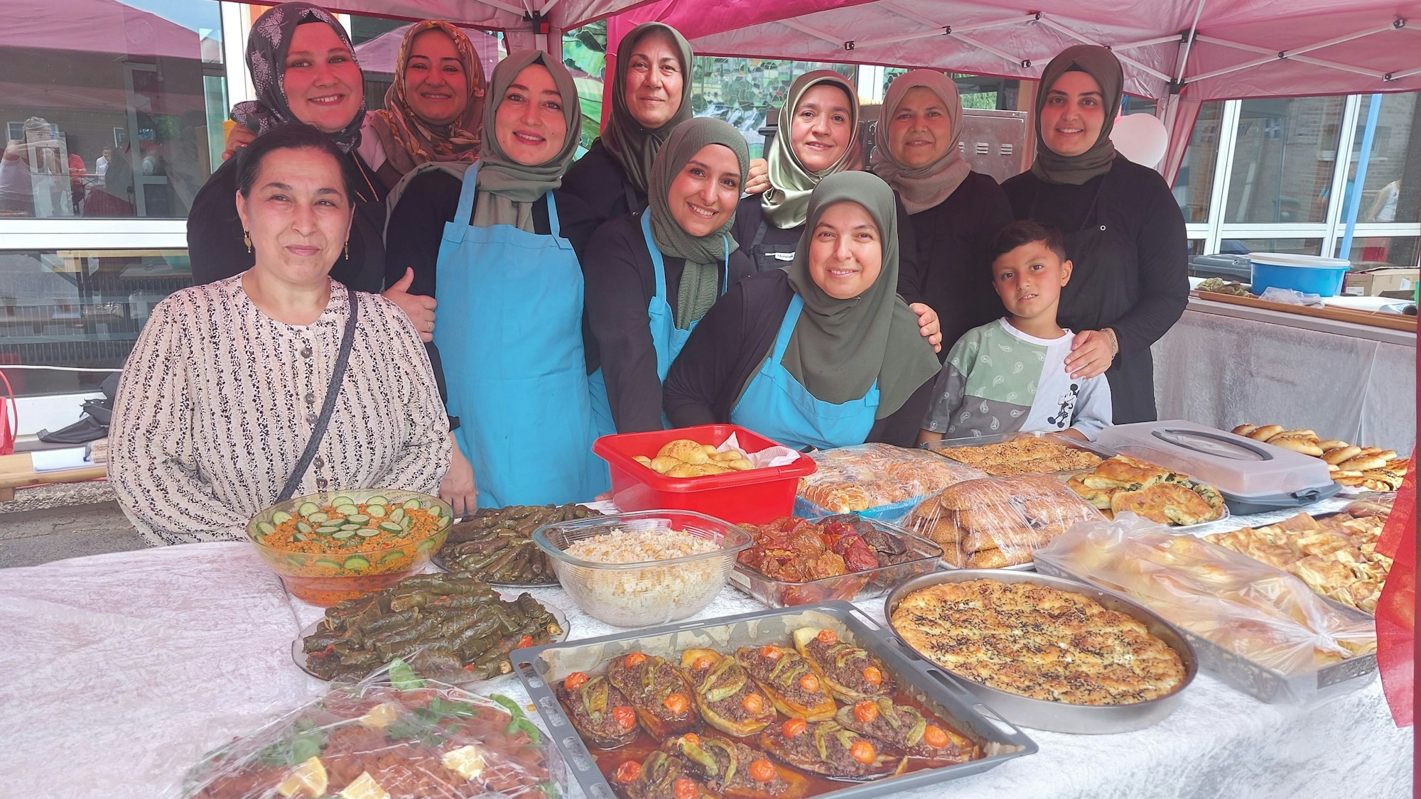 Lächelnde Frauen, fast alle mit Kopftüchern, stehen hinter einem großen Tisch, auf dem Platten und Schüsseln voller Essen stehen.