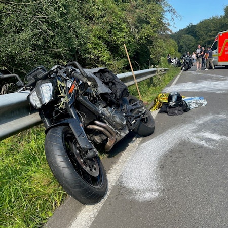 Ein beschädigtes Motorrad lehnt an der Leitplanke einer Straße, weiße Spuren sind auf dem Asphalt. Motorradkleidung und ein Helm liegen auf der Straße. Polizisten und ein Rettungswagen stehen im Hintergrund.
