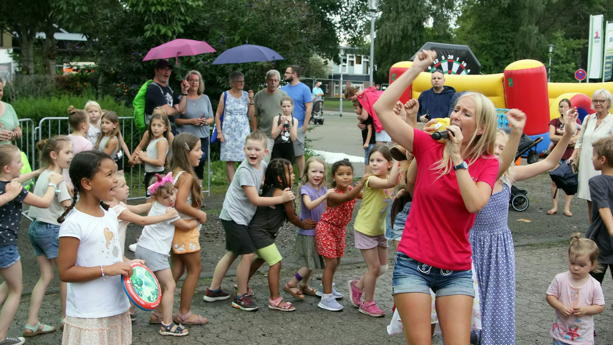 Eine Frau mit einem Mikrofon singt und tanzt, Kinder folgen ihr lachend in einer langen Polonaise.