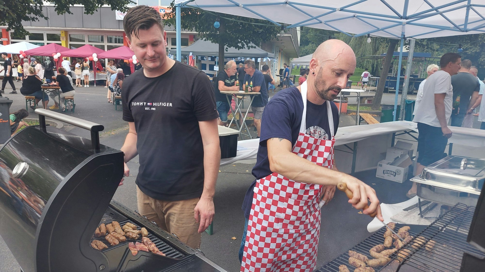 Zwei Männer, einer mit rot-karierter Schürze, braten Cevapcici auf zwei Grills.
